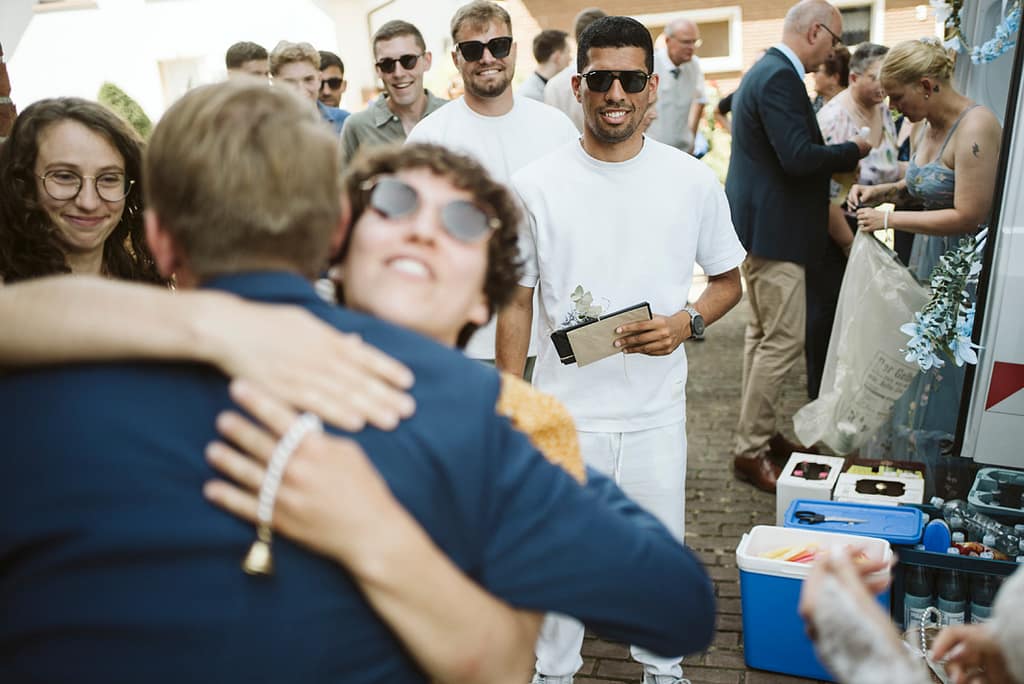 Eine Fru umarmt den Bräutigam. Im Hintergrund sieht man eine gruppe junger Männer, die alle eine Sonnenbrille tragen und ählich gekleidet sind. Alle strahlen.