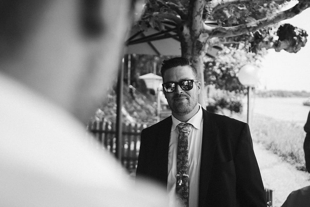 Schwarzweißaufnahme eines Gastes, der eine prägnante Sonnenbrille trägt. Fotografiert über die Schulter des Gegenübers inmitten der Konversation.