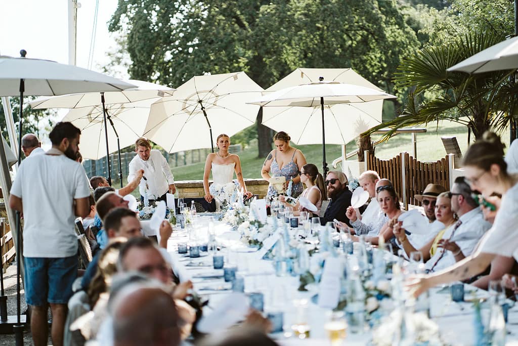 Gäste sitzen am großen, gedeckten Tisch. Das Brautpaar macht sich bereit mit Worten das Buffet zu eröffnen.