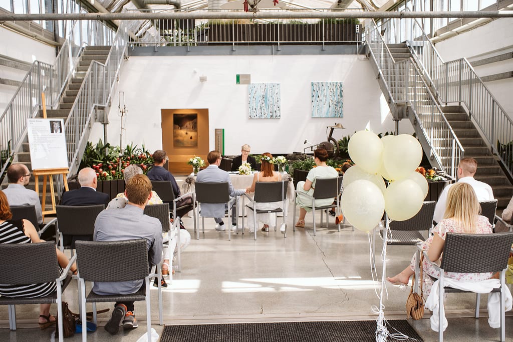 Der Saal im Innern des Elefanten. Der Tisch mit der Standesbeamtin, dem Brautpaar und den Trauzeugen ist in der Tiefe des Raumes zu sehen. Davor sieht man die Reihen der Gäste. Sehr auffallend sind einige weiße Ballons mit Helium gefüllt und an Schnüren. Die Ballons schweben ungefähr einen Meter über dem Boden, rechts im Bild. An der letzten sichtbaren Stuhlreihe.