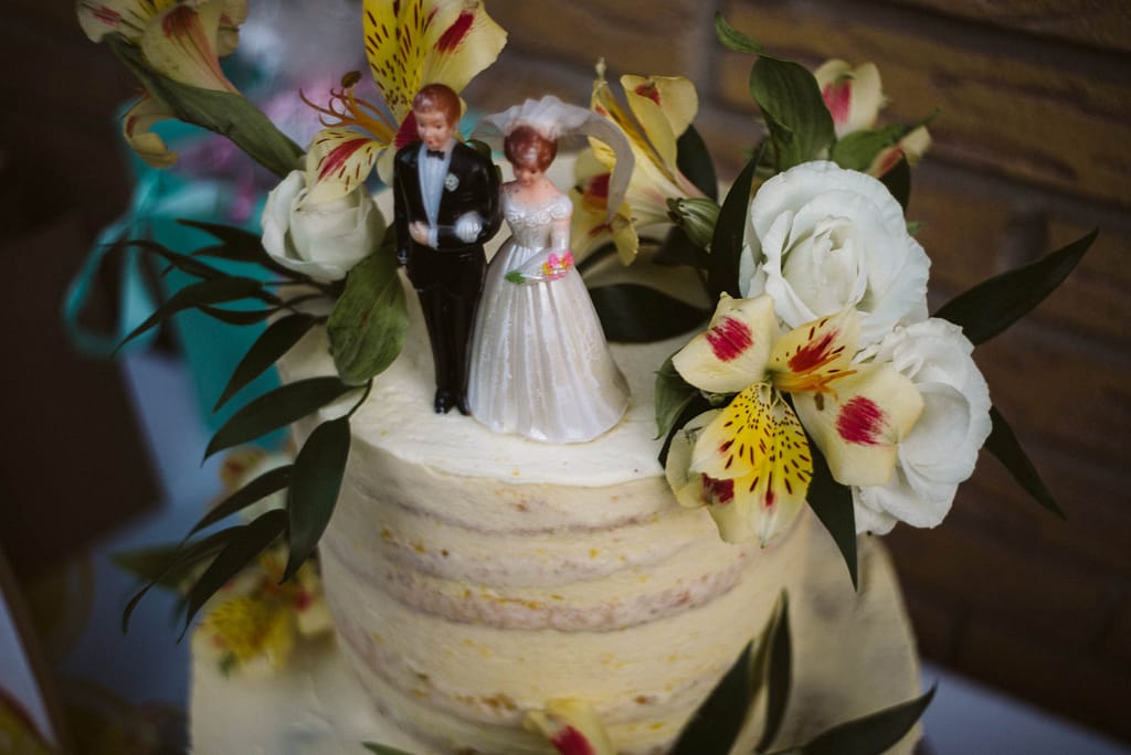 Hochzeitstorte mit zwei Figuren, Braut und Bräutigam in klassischen Outfits. Die Torte ist in Gelbtönen gestaltet. Dekoration sind gelbliche Blumen.