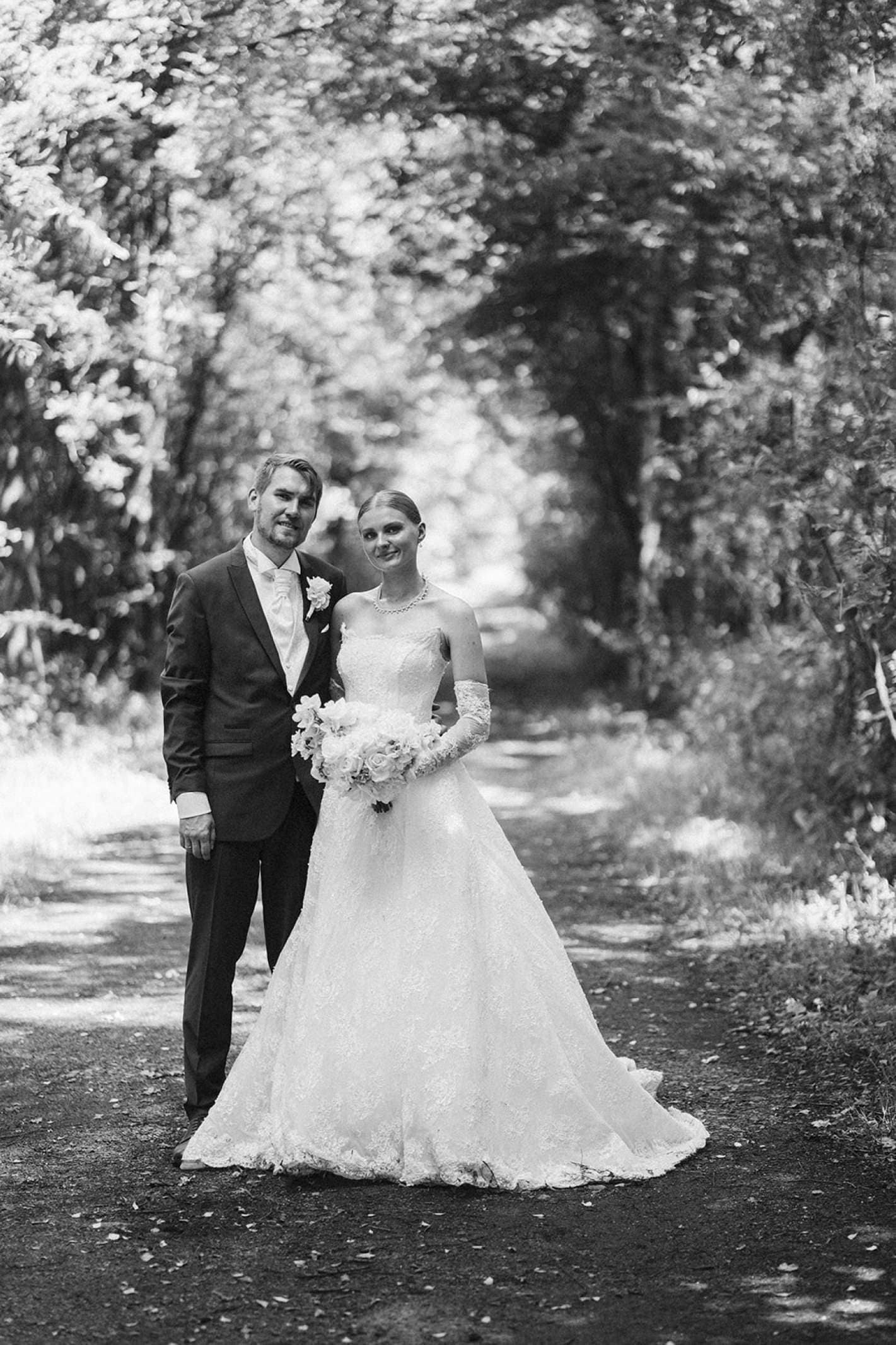 Das Brautpaar auf einem Waldweg. Klassisches Hochzeitsfoto.Schwarzweiß. Wenig Tiefenschärfe. Beide umarmen sich und schauen sich in die Augen.