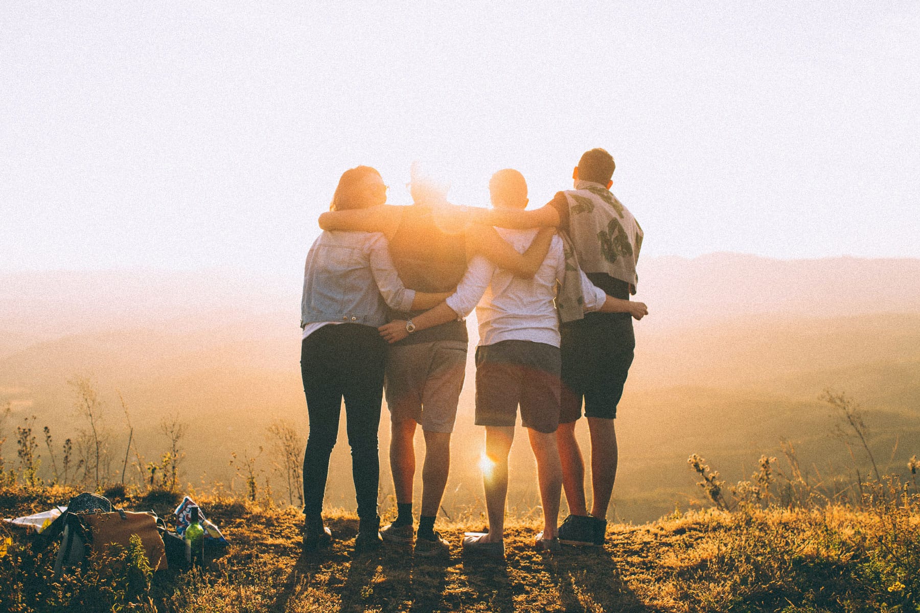 sugatatyler-gutschein-friends-04 Vier Freunde im Gegenlicht mit den Rücken zur Kamera gerichtet. Sie halten sich fest und schauen in den Sonnenaufgang